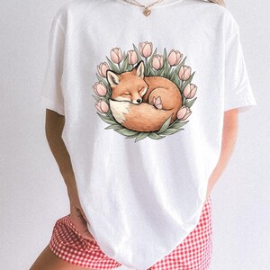 Peut inclure: Un t-shirt blanc avec une illustration graphique centrale représentant un renard orange endormi, enroulé à l'intérieur d'une couronne circulaire de tulipes roses et de feuilles vertes, avec un petit papillon posé sur le dos du renard.
