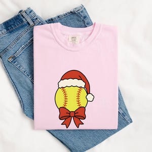 May include: A light pink short-sleeve t-shirt featuring a graphic of a yellow softball wearing a red Santa hat with a white trim and a red bow at the bottom, paired with a pair of blue denim jeans with a classic five-pocket design and metal rivets.