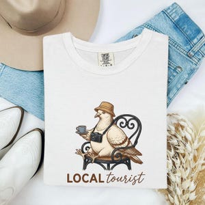 May include: White t-shirt featuring a cartoon pigeon in a hat, holding a camera and coffee, seated in a wrought-iron chair. The text "LOCAL tourist" is printed below the image.