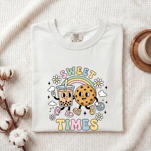 May include: A white t-shirt featuring a graphic print of a bubble tea cup and a chocolate chip cookie with arms and legs. The design includes a rainbow, daisies, and the text "SWEET TIMES" in pastel colors. The shirt has a crew neck and short sleeves.