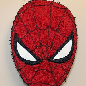 May include: A red and black Spider-Man mask pi&ntilde;ata. The mask features a red webbed design with black outlines and white eye shapes. The pi&ntilde;ata is hanging from a white string.