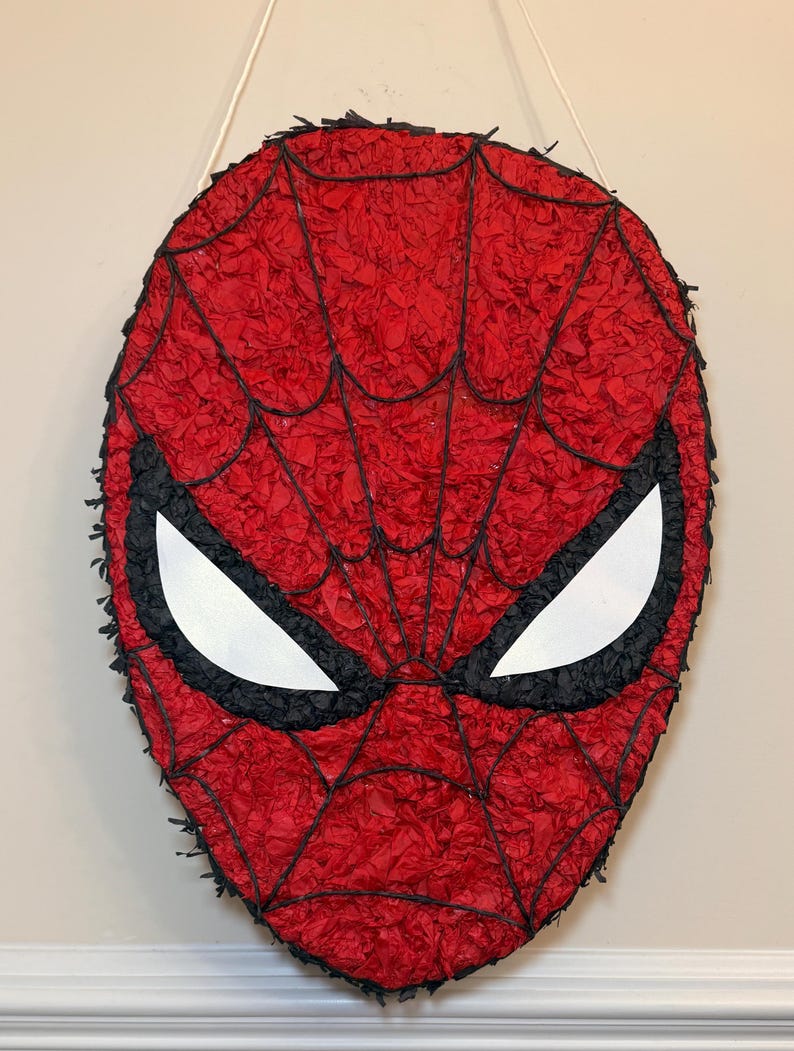 May include: A red and black Spider-Man mask pi&ntilde;ata. The mask features a red webbed design with black outlines and white eye shapes. The pi&ntilde;ata is hanging from a white string.
