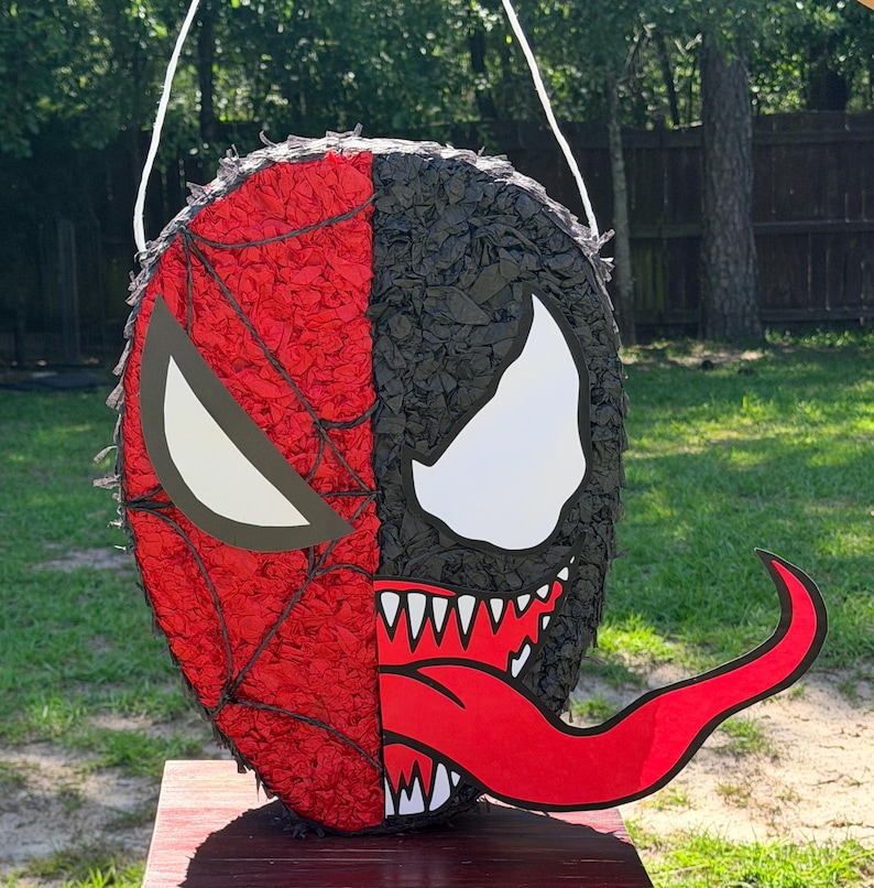 May include: A round pi&ntilde;ata featuring a split design with a red Spider-Man side displaying a black web pattern and a white eye, and a black Venom side showing a large white eye, sharp white teeth, and a long, protruding red tongue.