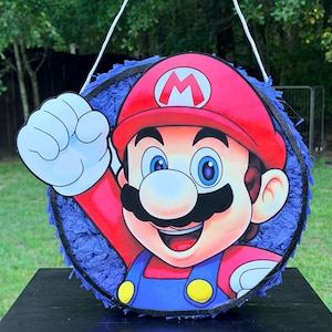 May include: A round pinata featuring a graphic of Mario with a red hat, blue overalls, and a white glove raised in a fist. The circular frame is decorated with blue fringed paper and a black border, suspended by a white string for hanging.
