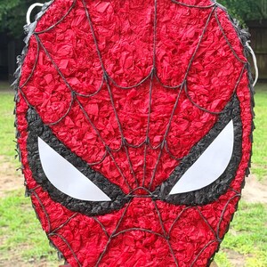 May include: A red and black Spider-Man pi&ntilde;ata. The mask-shaped pi&ntilde;ata features a red background with a black web design and white eye shapes. The pi&ntilde;ata is made of red tissue paper.