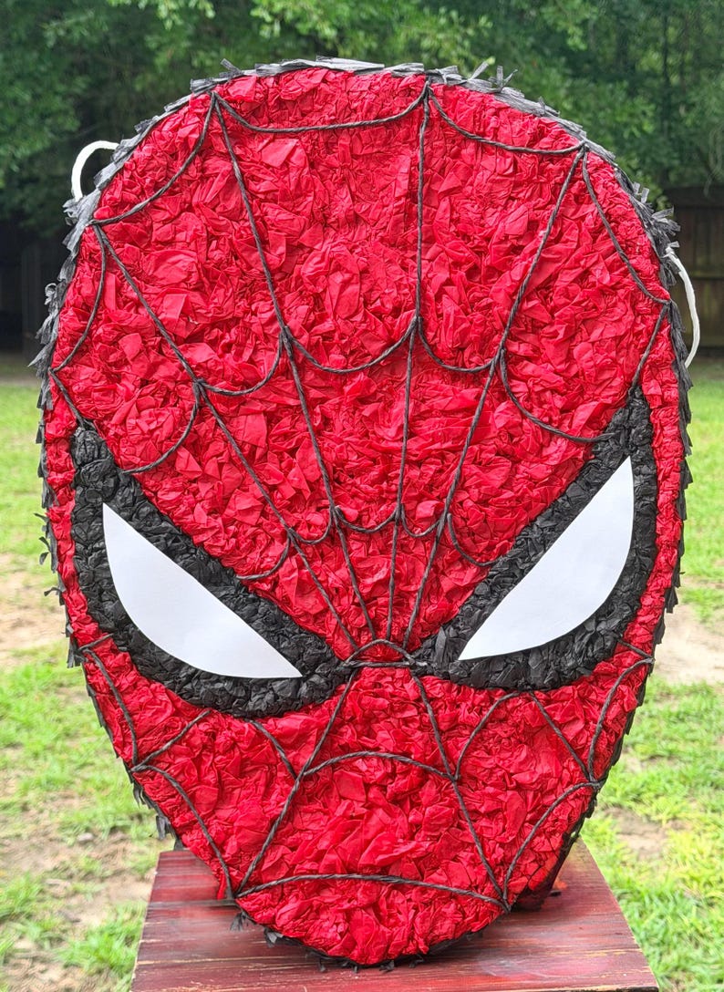 May include: A red and black Spider-Man pi&ntilde;ata. The mask-shaped pi&ntilde;ata features a red background with a black web design and white eye shapes. The pi&ntilde;ata is made of red tissue paper.