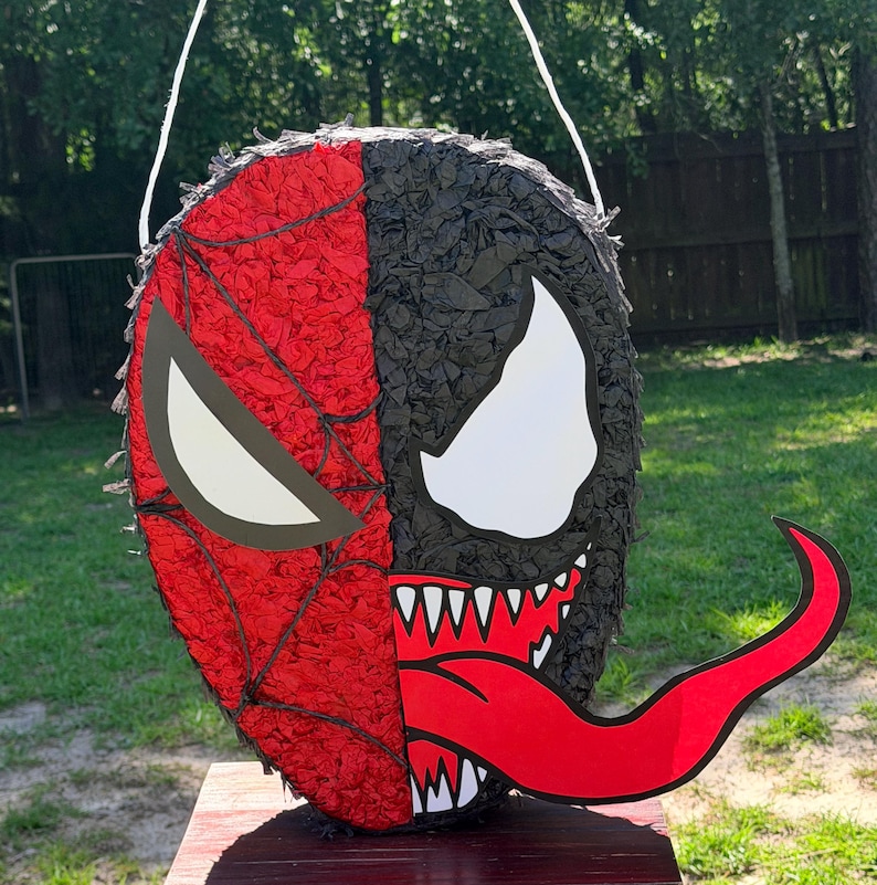 May include: A round pi&ntilde;ata featuring a split design. The left side is red with a black web pattern and a white eye shape. The right side is black with a white eye shape, a wide mouth with sharp white teeth, and a long, protruding red tongue.