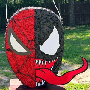 May include: A round pi&ntilde;ata featuring a split design with a red spiderweb pattern on the left side and a black textured surface with a large white eye and a long red tongue on the right side. The pi&ntilde;ata includes a white string for hanging.