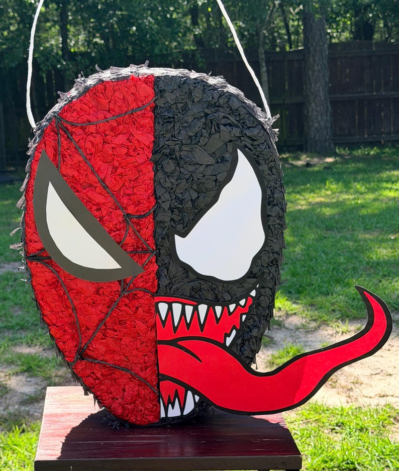 May include: A round pi&ntilde;ata featuring a split design with a red spiderweb pattern on the left side and a black textured surface with a large white eye and a long red tongue on the right side. The pi&ntilde;ata includes a white string for hanging.