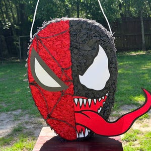 May include: A round pi&ntilde;ata featuring a split-face design. The left side is red with a black web pattern and a white eye shape. The right side is black with a white eye shape, sharp white teeth, and a long, curved red tongue extending outward.