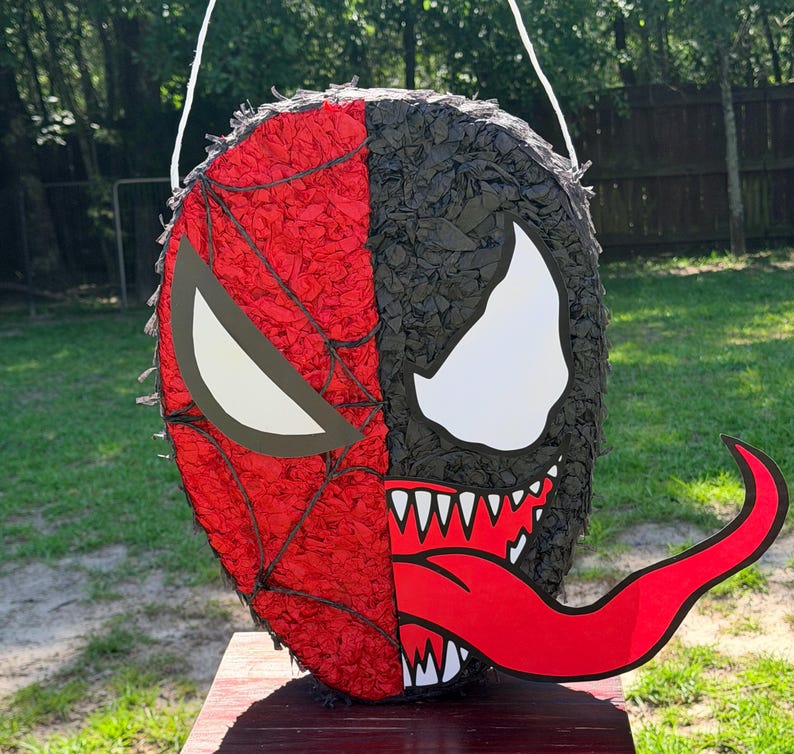 May include: A round pi&ntilde;ata featuring a split-face design. The left side is red with a black web pattern and a white eye shape. The right side is black with a white eye shape, sharp white teeth, and a long, curved red tongue extending outward.