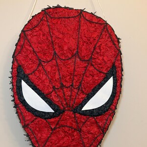 May include: A red Spider-Man mask pi&ntilde;ata with black webbing and eye outlines. The mask is made of red crepe paper and has white eye shapes. A white string is attached for hanging.