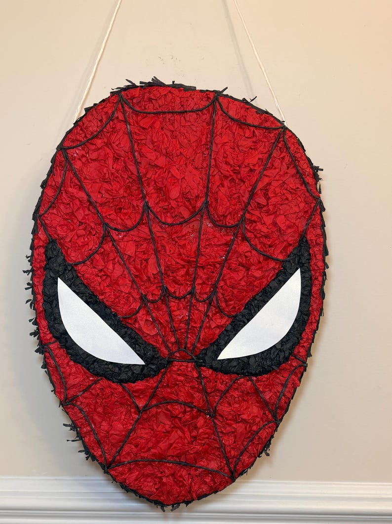 May include: A red Spider-Man mask pi&ntilde;ata with black webbing and eye outlines. The mask is made of red crepe paper and has white eye shapes. A white string is attached for hanging.