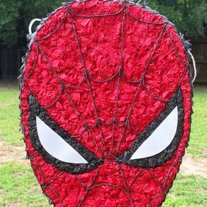 May include: A red Spider-Man mask pi&ntilde;ata with black webbing and eye details. The mask is made of red tissue paper and has white eye shapes. The pi&ntilde;ata is designed to resemble the superhero's face.