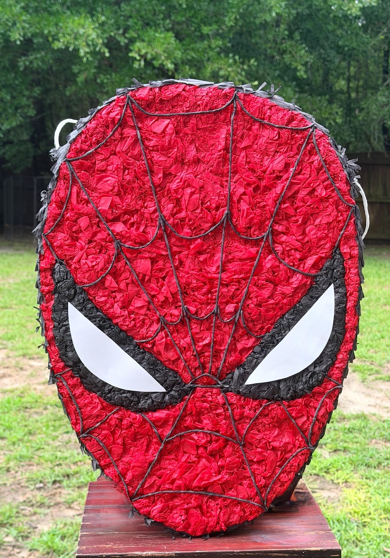 May include: A red Spider-Man mask pi&ntilde;ata with black webbing and eye details. The mask is made of red tissue paper and has white eye shapes. The pi&ntilde;ata is designed to resemble the superhero's face.