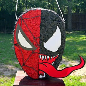 May include: A round pi&ntilde;ata featuring a split design. The left side is red with a black web pattern and a white eye. The right side is black with a large white eye, sharp white teeth, and a long, protruding red tongue. A white string is attached.