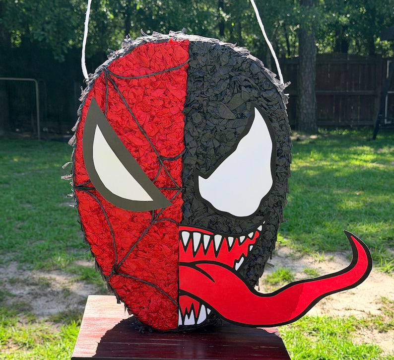 May include: A round pi&ntilde;ata featuring a split design. The left side is red with a black web pattern and a white eye. The right side is black with a large white eye, sharp white teeth, and a long, protruding red tongue. A white string is attached.