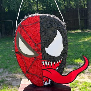 May include: A round pi&ntilde;ata featuring a split design with a red spider-web pattern on one side and a black textured surface with a large white eye and a long red tongue on the other side. The object includes a white string loop for hanging purposes.
