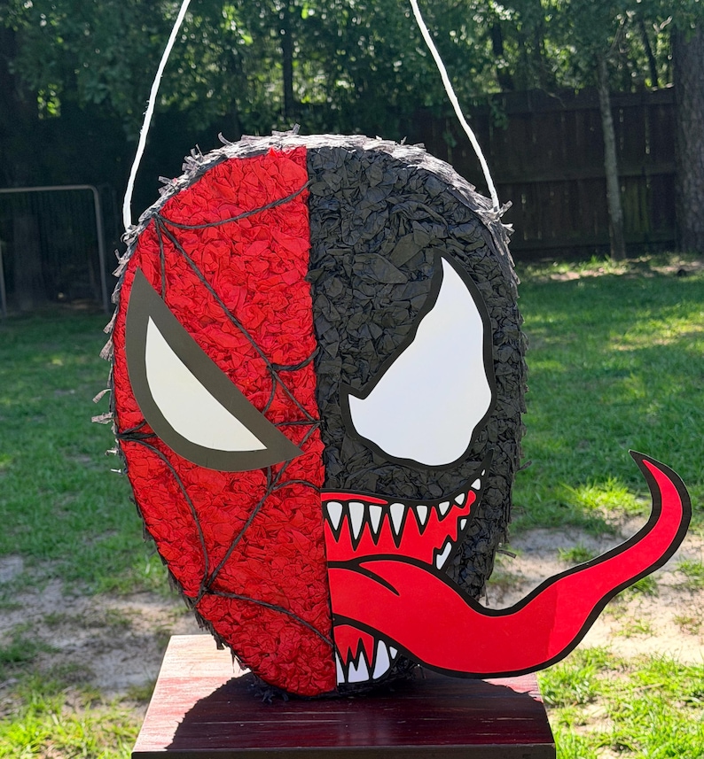 May include: A round pi&ntilde;ata featuring a split design with a red spider-web pattern on one side and a black textured surface with a large white eye and a long red tongue on the other side. The object includes a white string loop for hanging purposes.