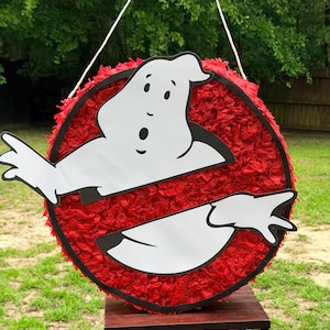 May include: A round piñata featuring a red circular background made of fringed paper and a white ghost character with black outlines. The ghost is positioned across the center of the red circle, with its arms extending outward to the sides.