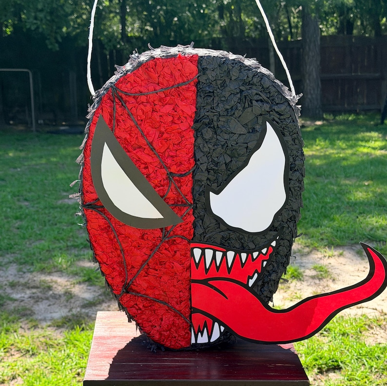 May include: A round pi&ntilde;ata featuring a split design. The left side is red with a black web pattern and a white eye shape. The right side is black with a large white eye, sharp white teeth, and a long, protruding red tongue extending to the right.