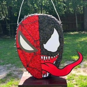 May include: A round pi&ntilde;ata featuring a split-face design. The left side is red with a black web pattern and a white eye, while the right side is black with a large white eye, sharp white teeth, and a long, curved red tongue extending outwards.