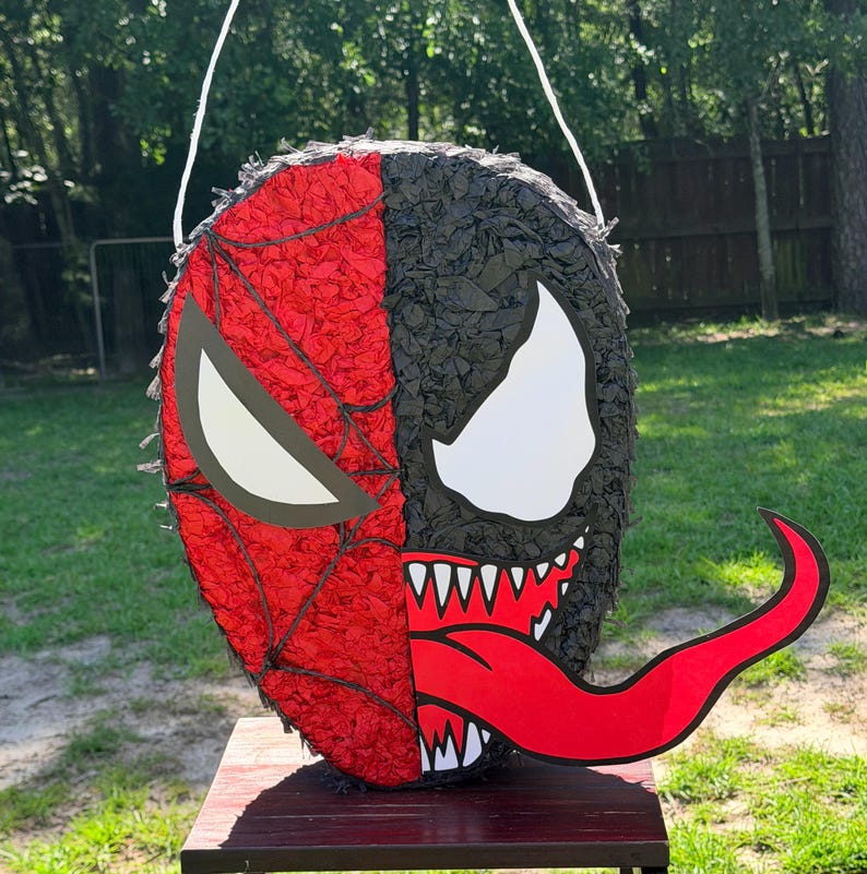 May include: A round pi&ntilde;ata featuring a split-face design. The left side is red with a black web pattern and a white eye, while the right side is black with a large white eye, sharp white teeth, and a long, curved red tongue extending outwards.