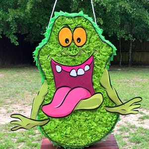 May include: A green piñata shaped like a cartoon creature with a wide open mouth, a long pink tongue, and two large yellow eyes. The body is covered in layers of green tissue paper fringe, and it features two light green arms extending outward.