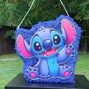 May include: A blue piñata featuring a large, colorful graphic of the character Stitch from Lilo and Stitch. The character is depicted with large dark eyes, a wide smile, and pink inner ears. The piñata is covered in dark blue fringe paper.