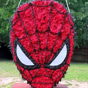 May include: A red and black Spider-Man pi&ntilde;ata, featuring white eye shapes and black webbing. The pi&ntilde;ata is made of red tissue paper and black trim, hanging from a white string. The pi&ntilde;ata is on a red table, with a green background.