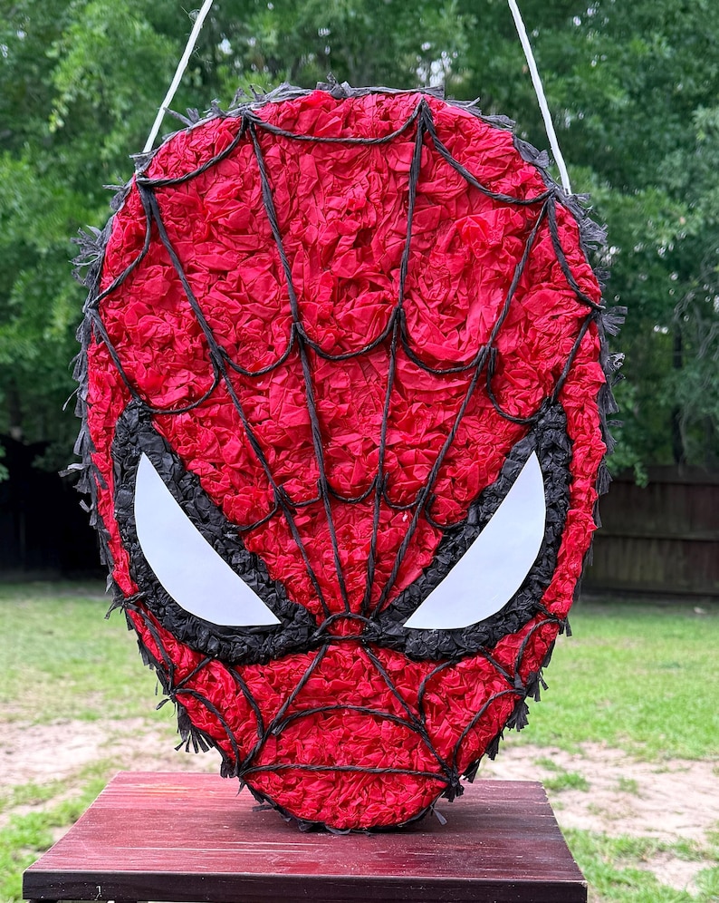 May include: A red and black Spider-Man pi&ntilde;ata, featuring white eye shapes and black webbing. The pi&ntilde;ata is made of red tissue paper and black trim, hanging from a white string. The pi&ntilde;ata is on a red table, with a green background.