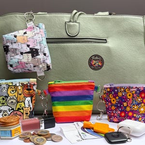 May include: A sage green tote bag featuring a zipped pocket and a circular logo patch. Four small zipped pouches are displayed in front: one with a cat print, one with a dog print, one with rainbow stripes, and one with a colourful floral pattern.