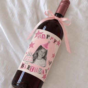 Puede incluir: Una botella de vino de cristal con tap&oacute;n de rosca rosa y un lazo rosa claro atado al cuello. La etiqueta muestra la foto de un ni&ntilde;o con un gorro de fiesta, un pastel de cumplea&ntilde;os, estrellas rosas brillantes y el texto HAPPY BIRTHDAY.