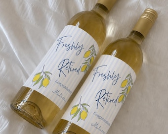 May include: Two glass wine bottles with gold screw caps, filled with a light yellow liquid. Each bottle features a white label with light blue vertical stripes, yellow lemon illustrations, and the text Freshly Retired Congratulations Melissa.