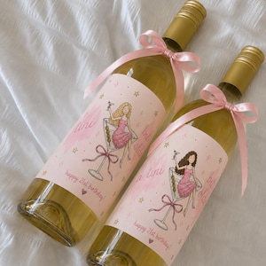 Puede incluir: Dos botellas de vino de cristal con tapones de rosca dorados, cada una adornada con un lazo de satén rosa claro. Las etiquetas muestran una ilustración de una persona en una copa de martini, con el texto 'a tini bit older' y 'happy 21st birthday!'