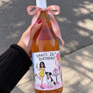 Puede incluir: Una botella de vidrio con líquido ámbar, tapón de rosca blanco y un lazo de satén rosa. La etiqueta muestra el texto DANI'S 26TH BIRTHDAY! junto a una ilustración de una persona con vestido amarillo, un perro y un pastel de cumpleaños.