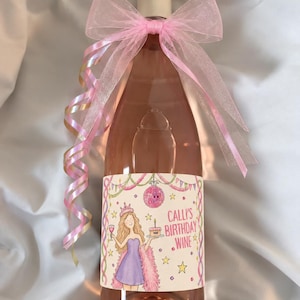 Puede incluir: Una botella de vino rosado adornada con un gran lazo de organza rosa translúcido y cintas rizadas. La etiqueta presenta una ilustración de una persona con vestido morado, una bola de discoteca, estrellas y el texto Calli's Birthday Wine.