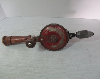 Puede incluir: Taladro manual antiguo con cuerpo de metal rojo y plateado, mango de madera y broca. El taladro muestra signos de desgaste, con pintura desconchada del metal. El mango es de color marrón claro. Una herramienta clásica para trabajos de madera y proyectos de bricolaje.