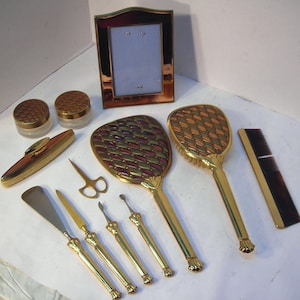 May include: A vintage vanity set with gold-toned metal handles and accents. The set includes two hand mirrors, a comb, a picture frame, two jars, a compact, scissors, and several manicure tools. The mirrors have a geometric pattern in shades of red, green, and gold.