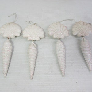 May include: Four white seashell ornaments with glitter accents. Each ornament features a scallop shell shape at the top and a spiraled conch shell shape below. The ornaments have a string for hanging.