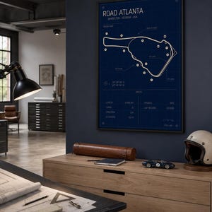 May include: A framed blueprint-style poster of the Road Atlanta racetrack in Braselton, Georgia, USA. The poster is in a black frame and features a white track map on a dark blue background. A wooden cabinet with a model race car, helmet, and leather case is in the foreground.