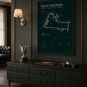 May include: A framed Mount Panorama race track map in dark green hangs above a dark wood and green leather cabinet. A silver trophy, a notebook, and a green race car model sit on the cabinet. A wall sconce with a cream shade is to the left.