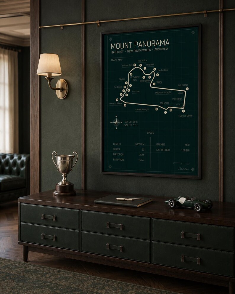 May include: A framed Mount Panorama race track map in dark green hangs above a dark wood and green leather cabinet. A silver trophy, a notebook, and a green race car model sit on the cabinet. A wall sconce with a cream shade is to the left.