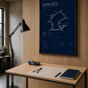 May include: A framed blue print of the Laguna Seca race track in Monterey, California, USA. The track map is displayed on a dark blue background with white lines and text. A small blue race car model sits on a wooden desk with a notebook and drafting tools.