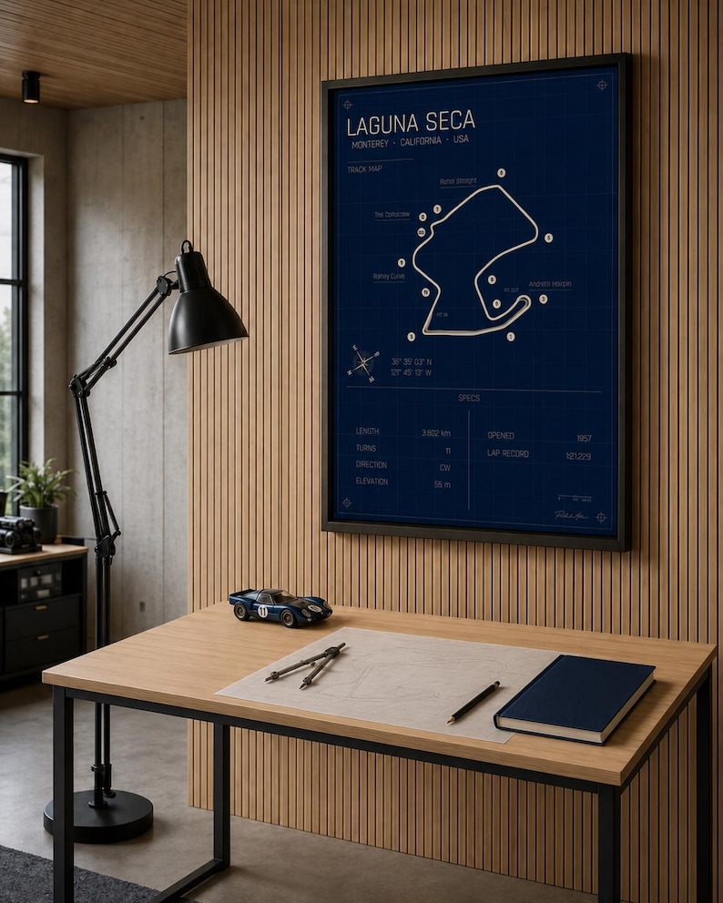 May include: A framed blue print of the Laguna Seca race track in Monterey, California, USA. The track map is displayed on a dark blue background with white lines and text. A small blue race car model sits on a wooden desk with a notebook and drafting tools.