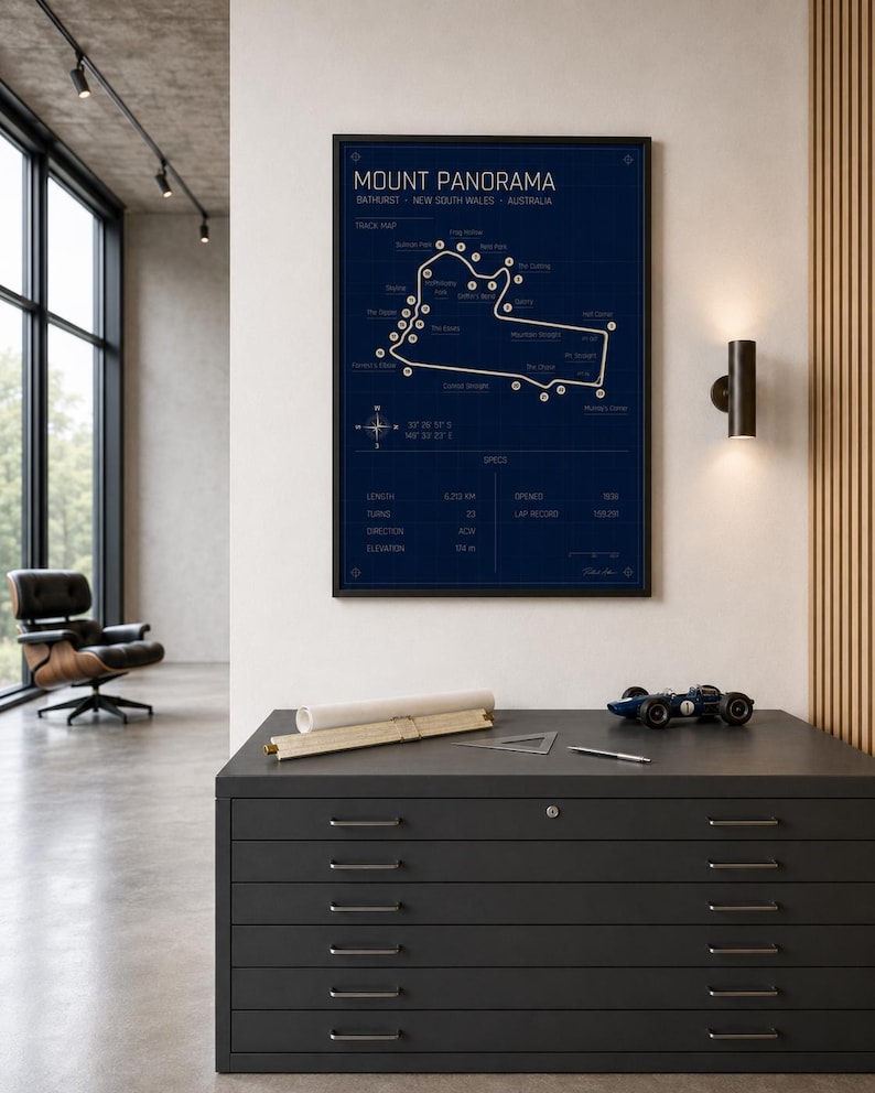 May include: A framed Mount Panorama race track map in navy blue with white detailing. The map is displayed above a dark gray cabinet with several drawers. A small blue race car model sits on top of the cabinet.