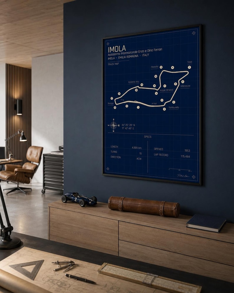 May include: A framed blueprint-style poster of the Imola race track in Italy, with white lines on a dark blue background. The poster includes track details and specifications. A model race car and a leather case sit on a wooden cabinet below.