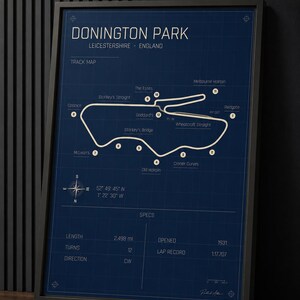 Op de afbeelding: Een ingelijste print in blauwdrukstijl van Donington Park, een racecircuit in Leicestershire, Engeland. De kaart van het circuit is wit op een donkerblauwe achtergrond, met gelabelde bochten en specificaties, waaronder een lengte van 4,02 km en een ronderecord van 1:17.707.