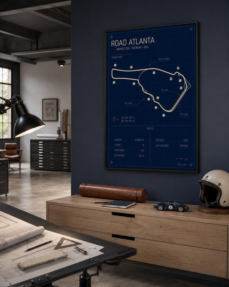 May include: A framed blueprint-style poster of the Road Atlanta racetrack in Braselton, Georgia, USA. The poster is in a black frame and features a white track map on a dark blue background. A wooden cabinet with a model race car, helmet, and leather case is in the foreground.