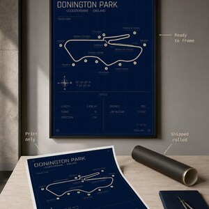 Op de afbeelding: Een ingelijste en opgerolde print van de Donington Park-circuitkaart in Leicestershire, Engeland. De kaart toont de witte circuitindeling op een donkerblauwe achtergrond, met details zoals lengte en aantal bochten.